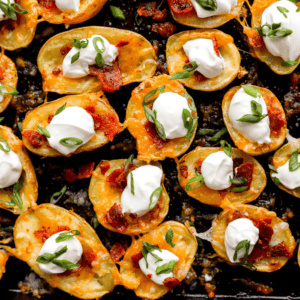 Close-up of golden baked potato skin bites topped with melted cheddar cheese, crispy bacon, sour cream, and sliced green onions on a dark baking tray.