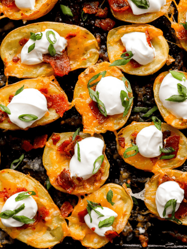 Close-up of golden baked potato skin bites topped with melted cheddar cheese, crispy bacon, sour cream, and sliced green onions on a dark baking tray.
