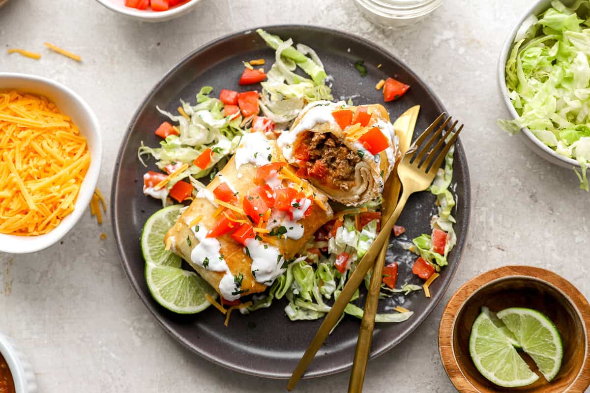 A sliced chimichanga showing the savory beef filling inside, served on a plate with lettuce, tomatoes, sour cream, and lime wedges, with a gold fork and knife.