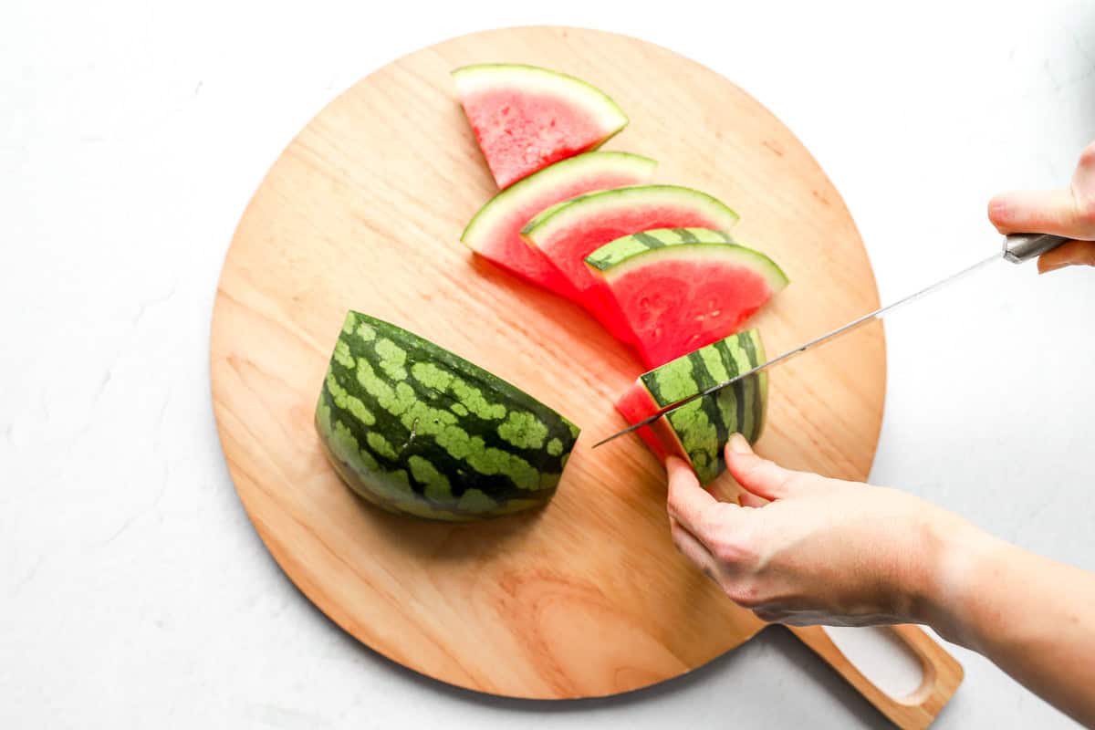 Knife slicing a watermelon half into thin triangular wedges on a wooden board.
