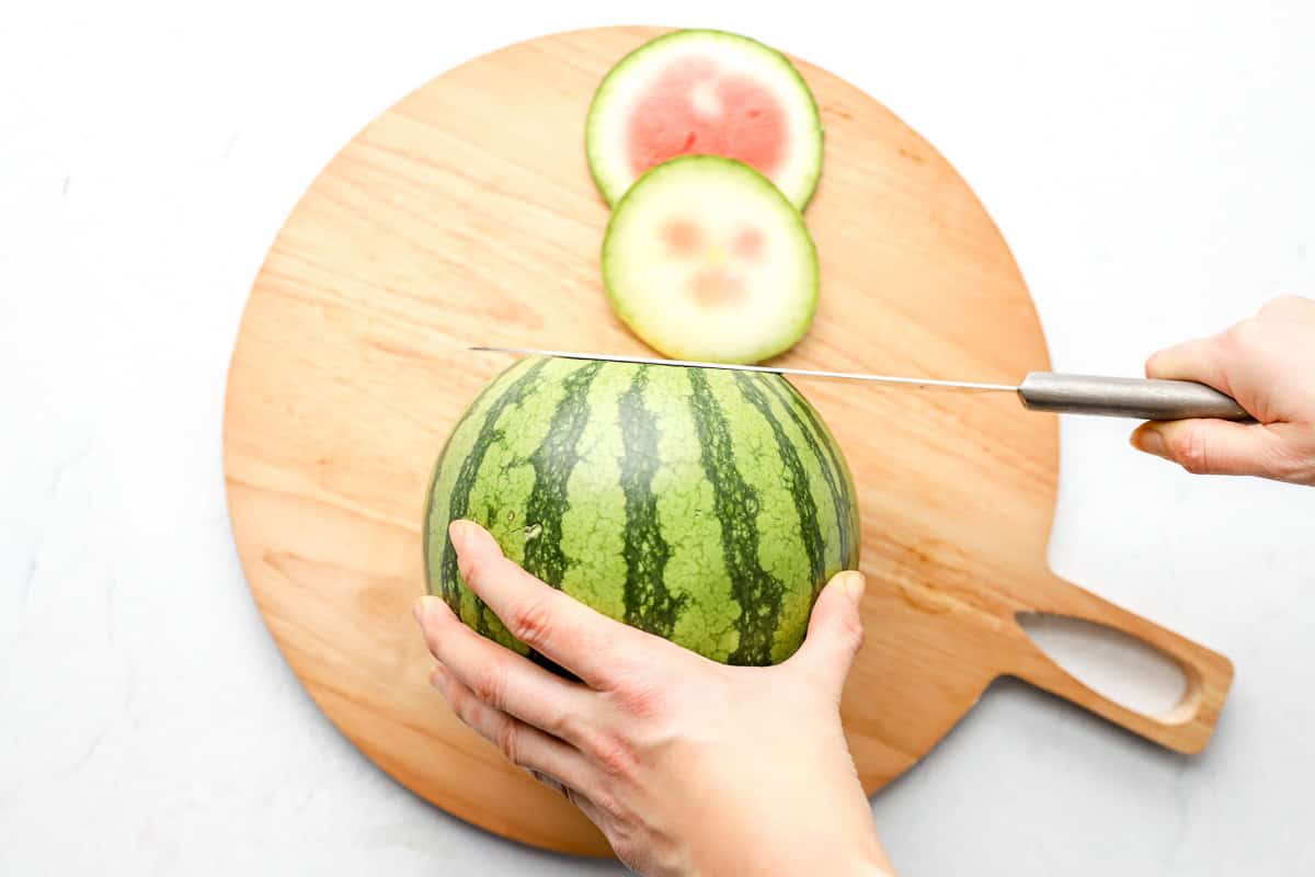 Hands holding a watermelon steady while slicing off the top end on a round wooden cutting board.