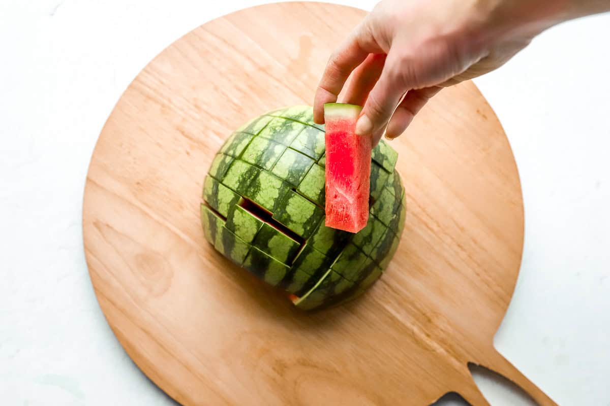 Hand lifting out a perfectly cut rectangular watermelon stick from the grid-cut half.