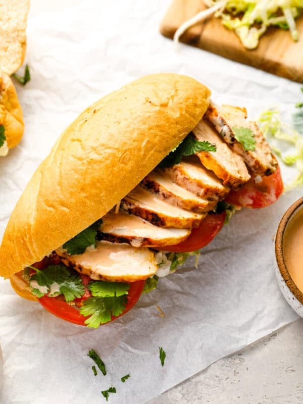 Close-up of a chicken torta sandwich on parchment paper, filled with sliced grilled chicken, tomato, lettuce, cilantro, and chipotle mayo on a bolillo roll.