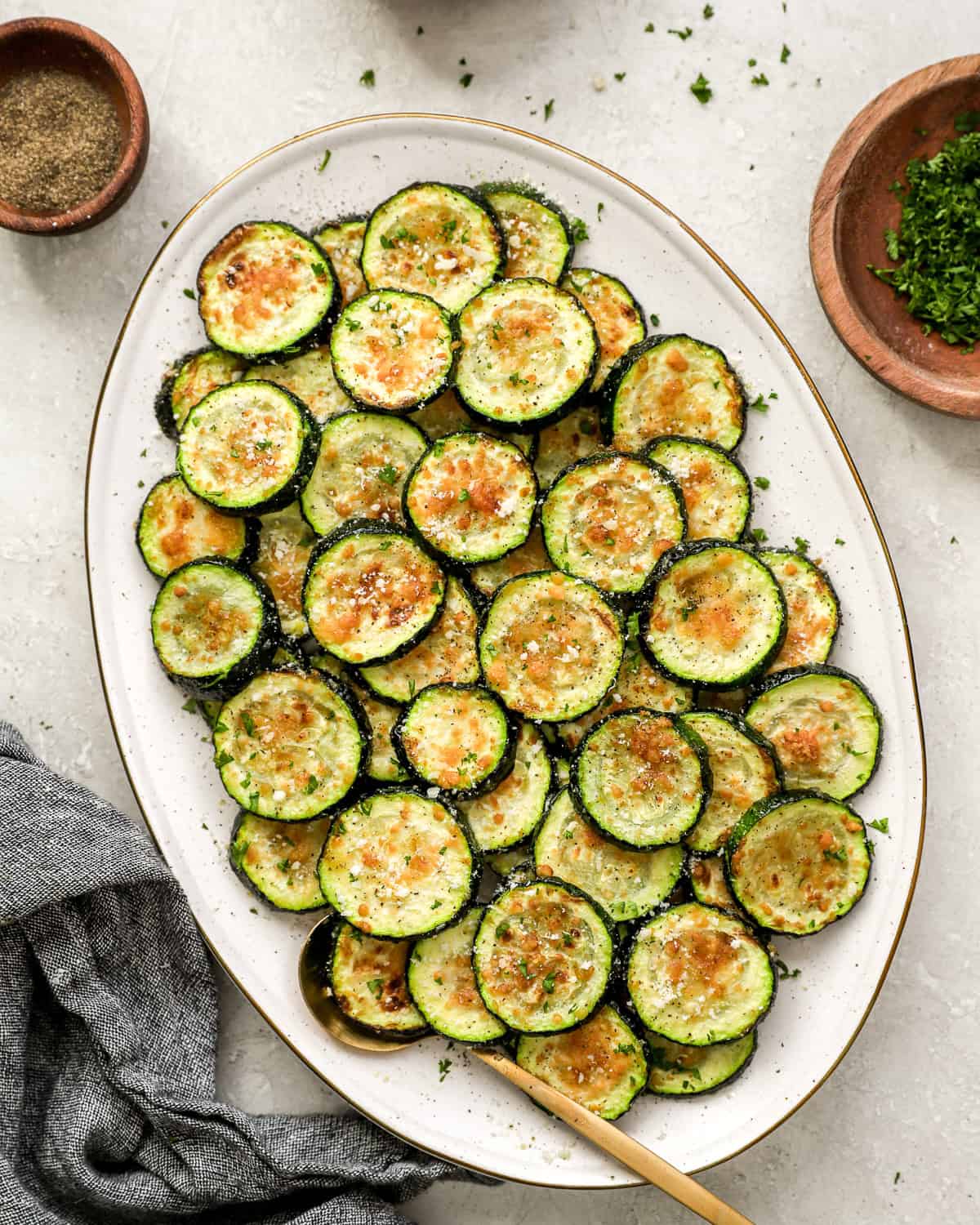 Oval platter of golden, crispy air fryer zucchini rounds sprinkled with Parmesan and chopped parsley, served with a gold spoon.