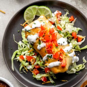 A golden, crispy chimichanga topped with diced tomatoes, shredded cheese, and a drizzle of sour cream, served on a bed of shredded lettuce with lime wedges.