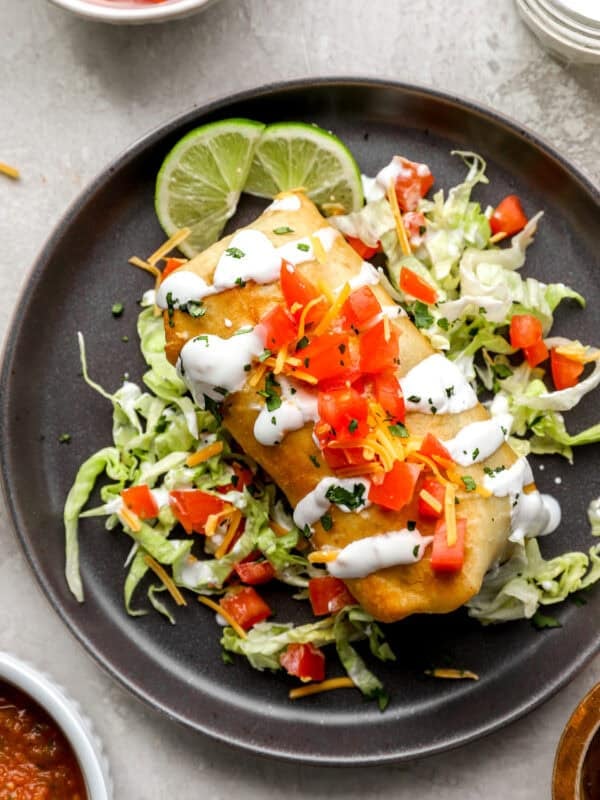 A golden, crispy chimichanga topped with diced tomatoes, shredded cheese, and a drizzle of sour cream, served on a bed of shredded lettuce with lime wedges.