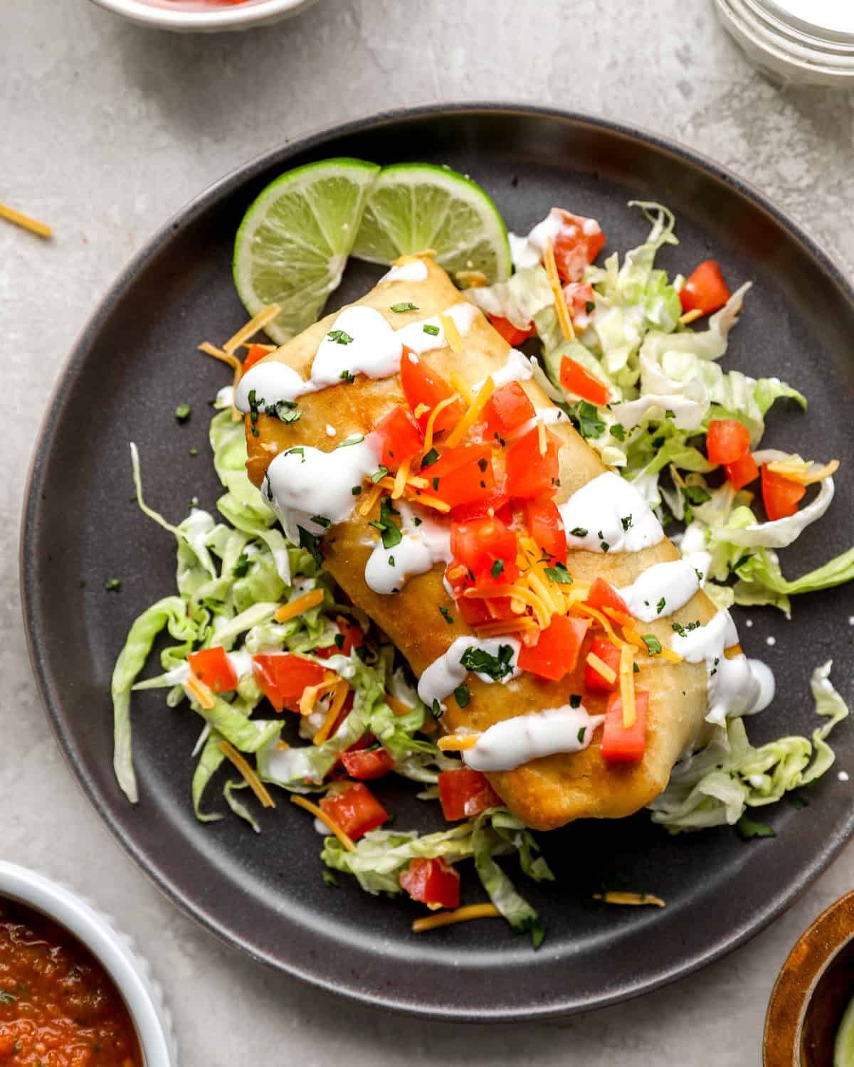 A golden, crispy chimichanga topped with diced tomatoes, shredded cheese, and a drizzle of sour cream, served on a bed of shredded lettuce with lime wedges.