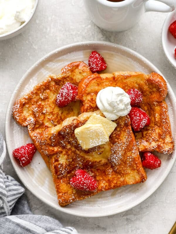 Overhead view of brioche French toast with butter, whipped cream, fresh raspberries, and maple syrup on a white plate.