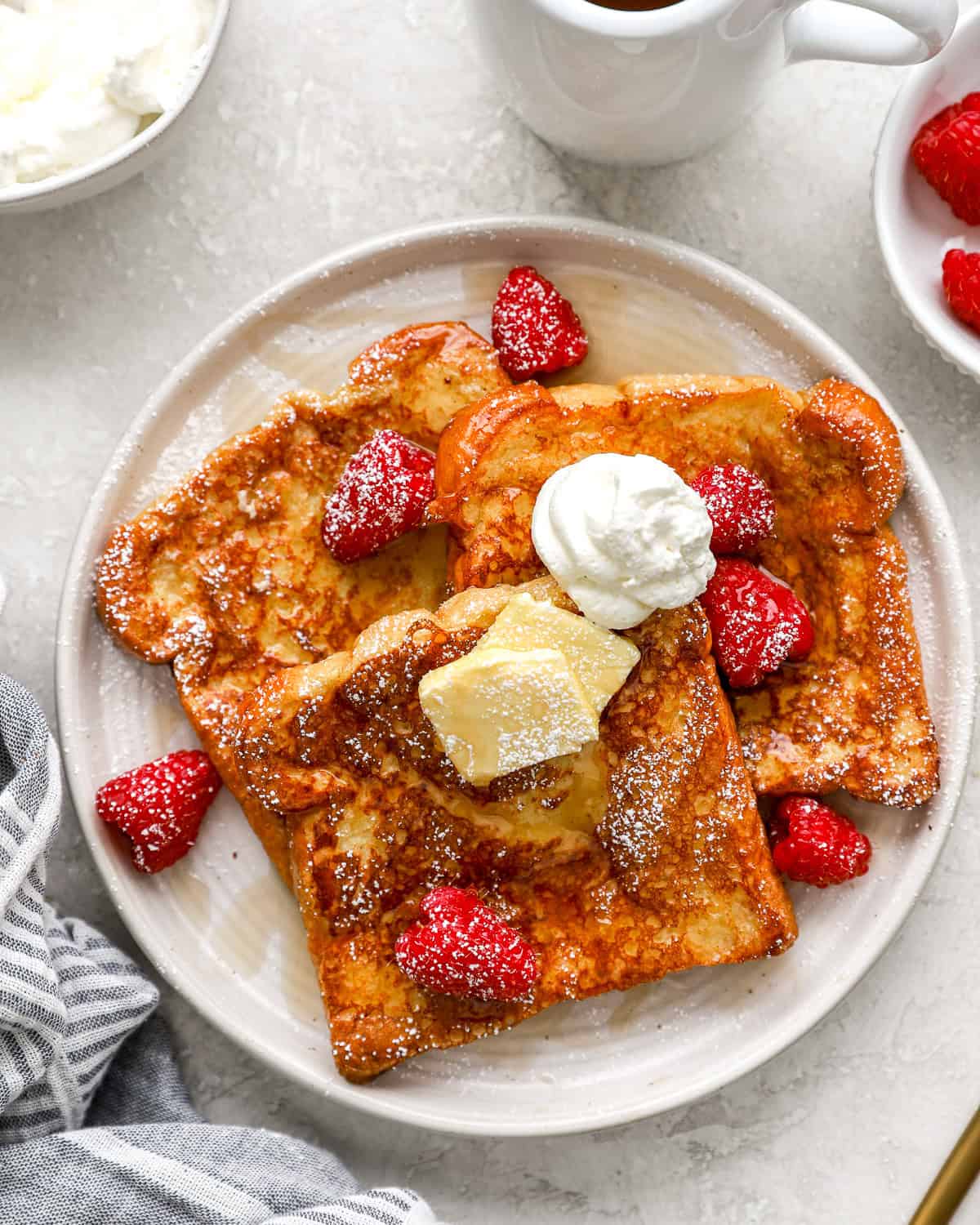 Overhead view of brioche French toast with butter, whipped cream, fresh raspberries, and maple syrup on a white plate.