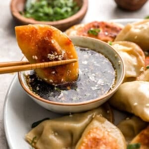 A golden-brown pan-fried chicken potsticker held by chopsticks being dipped into a soy-based dipping sauce topped with sesame seeds and chopped green onions.
