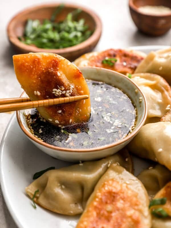 A golden-brown pan-fried chicken potsticker held by chopsticks being dipped into a soy-based dipping sauce topped with sesame seeds and chopped green onions.