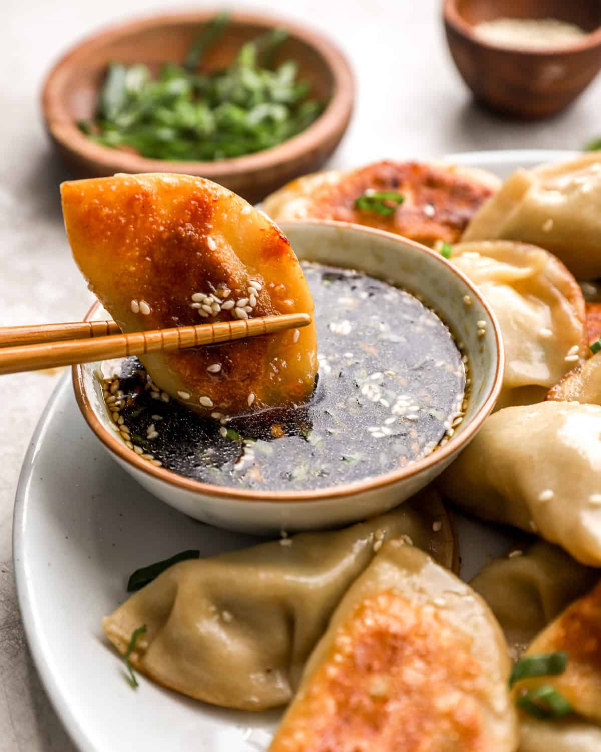 A golden-brown pan-fried chicken potsticker held by chopsticks being dipped into a soy-based dipping sauce topped with sesame seeds and chopped green onions.