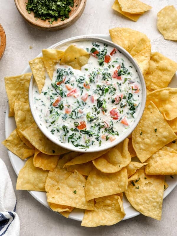 Bowl of creamy white cheese dip with spinach and tomatoes, surrounded by crispy tortilla chips on a white plate.