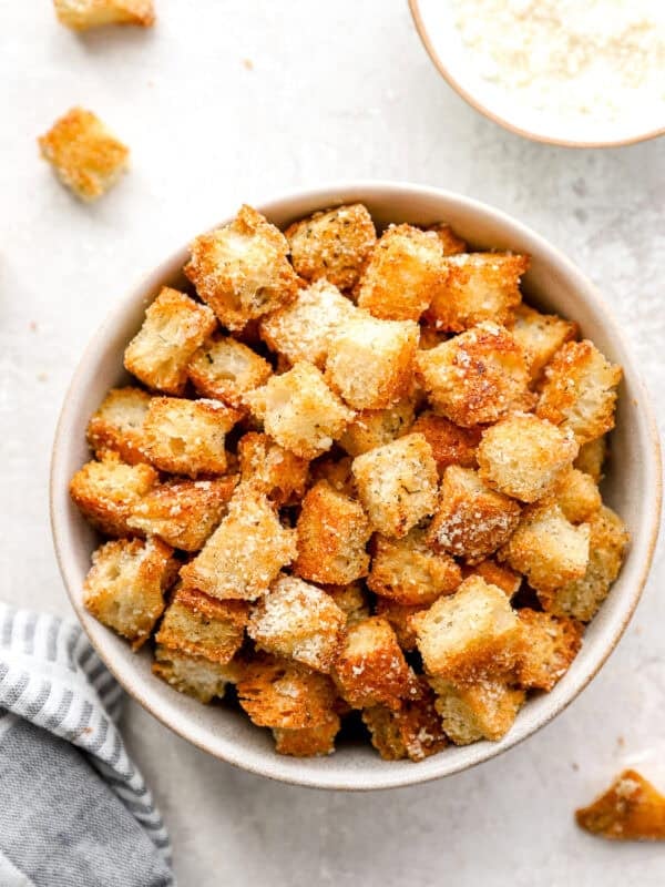 A bowl filled with golden, crispy homemade croutons, sprinkled with Parmesan cheese, on a light surface with a striped napkin beside it.