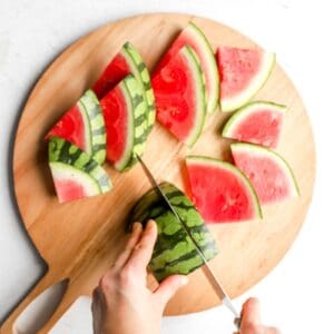 Sliced watermelon wedges arranged on a round wooden cutting board, with hands slicing a watermelon half into triangles.
