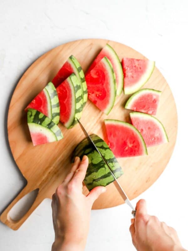 Sliced watermelon wedges arranged on a round wooden cutting board, with hands slicing a watermelon half into triangles.
