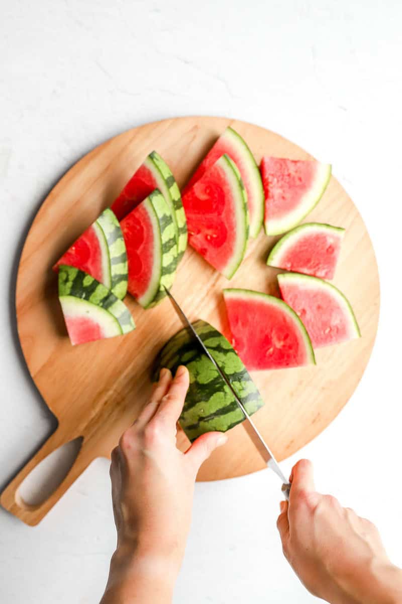 Sliced watermelon wedges arranged on a round wooden cutting board, with hands slicing a watermelon half into triangles.