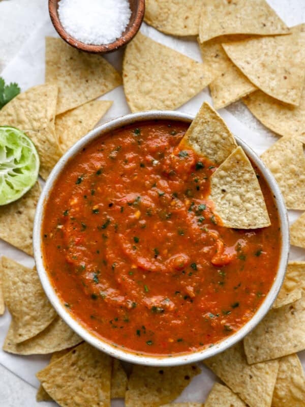 Bowl of salsa roja surrounded by tortilla chips, with a chip dipped into the salsa and garnishes like lime and sea salt on the side.