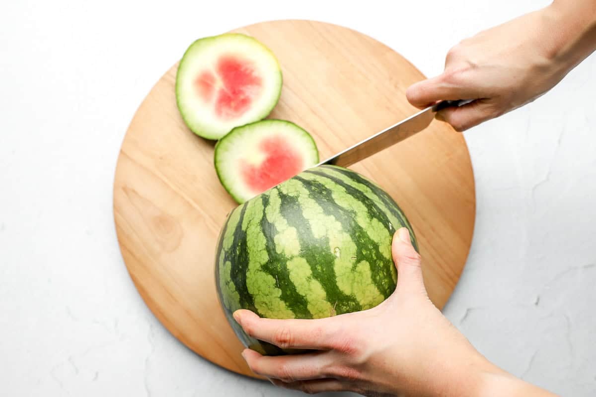 Hands slicing off the top end of a whole watermelon on a round wooden cutting board.