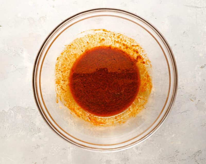 Overhead shot of a spice marinade in a glass bowl on a light surface, with bright orange-red color and seasoning residue on the sides.
