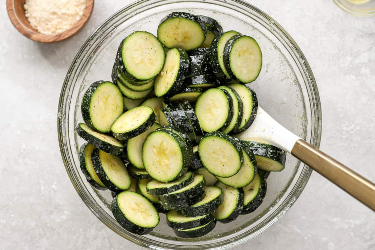 Glass bowl of raw zucchini slices tossed in olive oil and seasonings, ready for the air fryer, with grated Parmesan visible nearby.