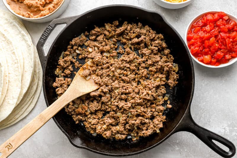 Seasoned ground beef with onions cooking in a cast iron skillet, surrounded by bowls of ingredients including tortillas, refried beans, shredded cheese, and diced tomatoes.