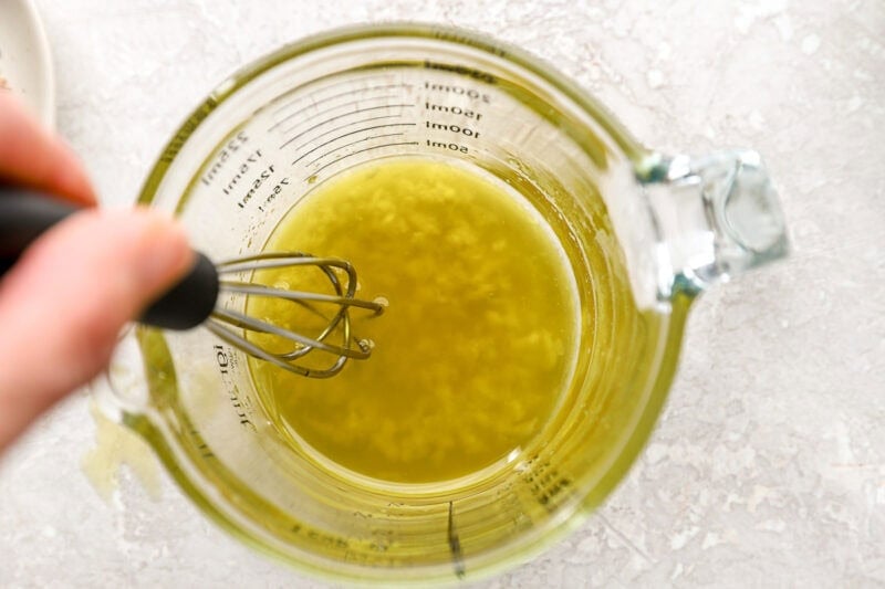 Close-up of minced garlic being whisked into olive oil in a glass measuring cup.