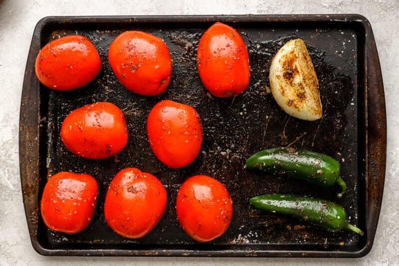 Raw roma tomatoes, onion, and jalapeños arranged on a baking sheet, seasoned and ready for roasting.