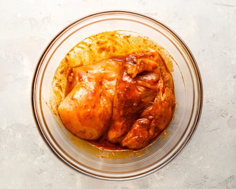 Raw chicken breasts marinating in a bowl of red spice marinade, fully coated and ready for cooking.