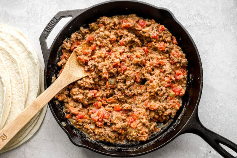 Ground beef filling mixed with refried beans and diced tomatoes simmering in a cast iron skillet, with a stack of tortillas beside it.