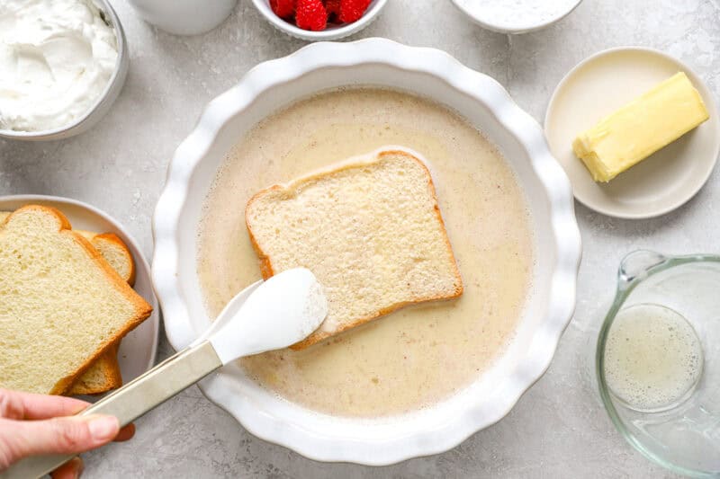 Brioche slice being dipped into custard mixture with tongs, surrounded by ingredients like butter, raspberries, and whipped cream.