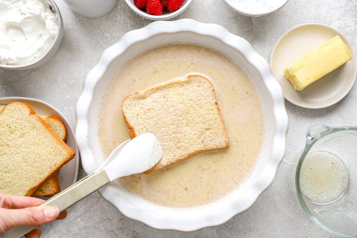 Brioche slice being dipped into custard mixture with tongs, surrounded by ingredients like butter, raspberries, and whipped cream.