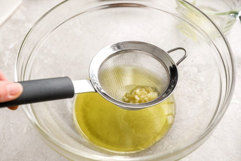 Minced garlic being strained from garlic-infused olive oil through a fine mesh sieve over a glass bowl.