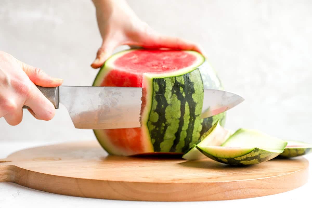 Close-up of a large knife removing the rind from the side of a standing watermelon.