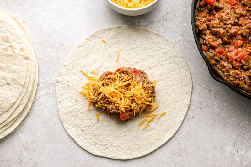 A flour tortilla topped with a scoop of beef filling and shredded cheese, ready to be wrapped.