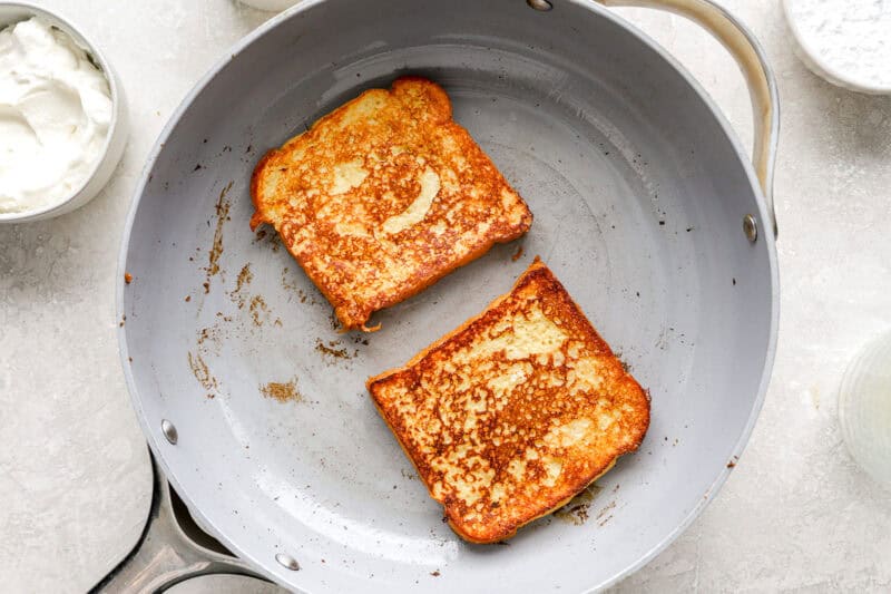 Two slices of brioche French toast cooking in a skillet, golden brown and crisping on the edges.