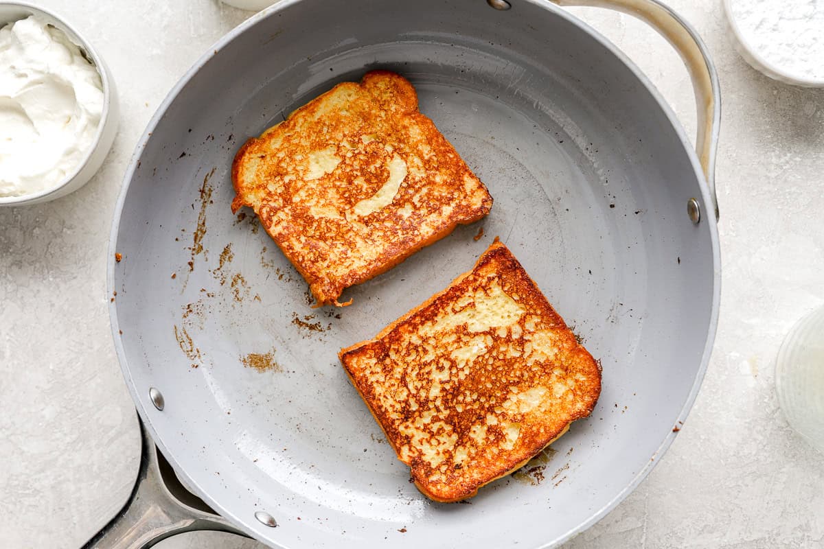 Two slices of brioche French toast cooking in a skillet, golden brown and crisping on the edges.