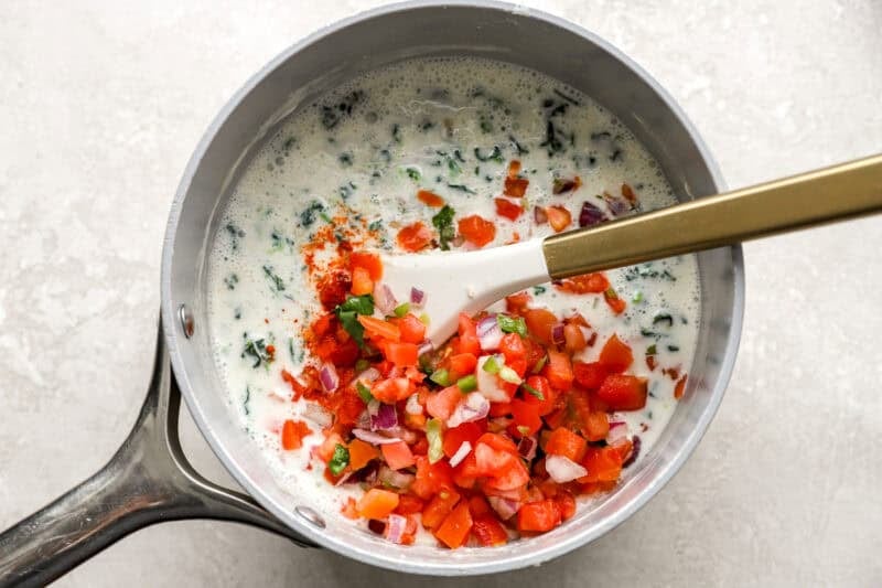 Fresh pico de gallo, spinach, and cayenne pepper being stirred into melted cheese and milk in a saucepan.