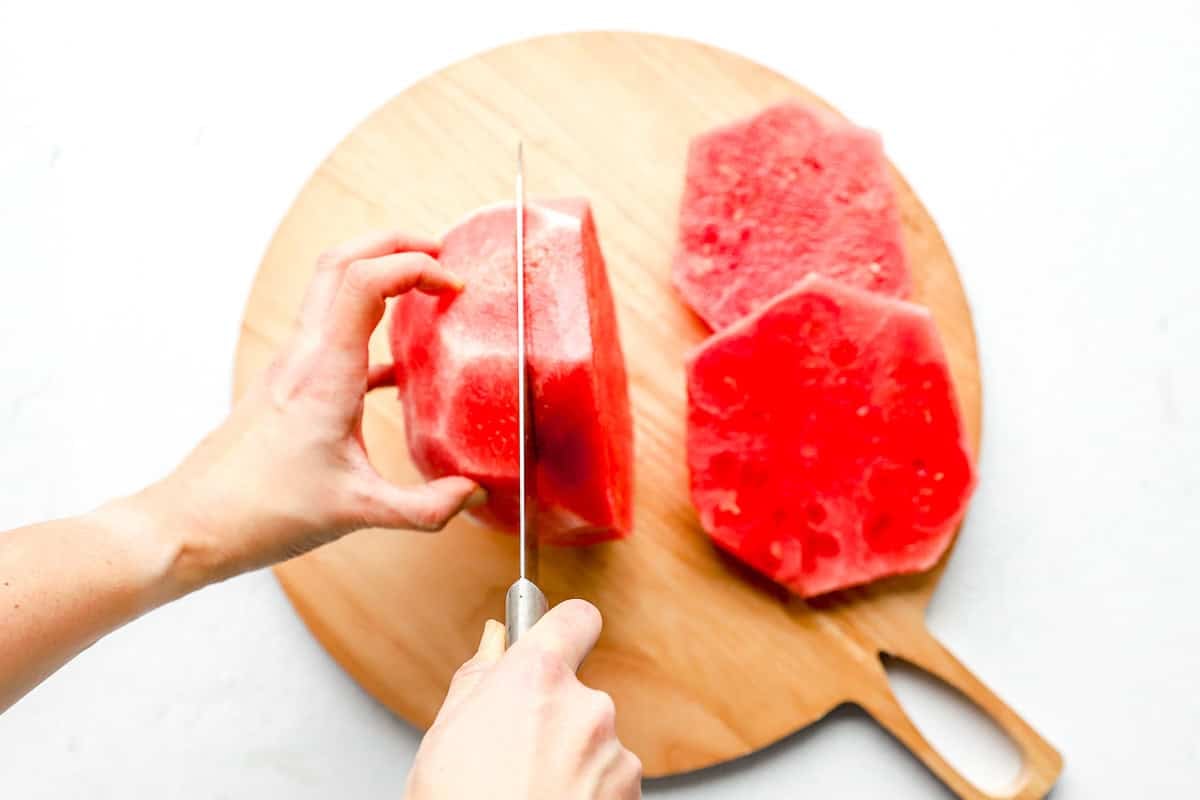 Knife slicing a peeled watermelon into thick round discs on a wooden cutting board.