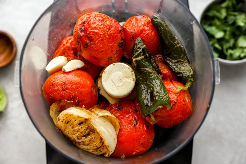 Roasted tomatoes, jalapeños, and onion placed in a food processor bowl with garlic cloves, ready to be blended.