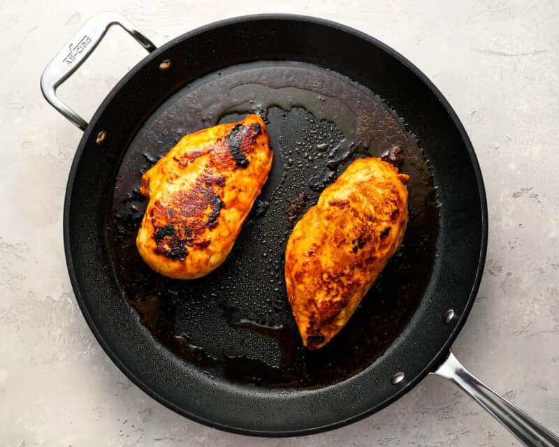 Two seasoned chicken breasts cooking in a black skillet, lightly charred and golden brown.