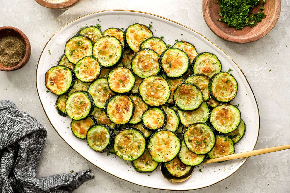 Finished air fryer zucchini on an oval platter, sprinkled with Parmesan and herbs, with a gold serving spoon and garnishes around.