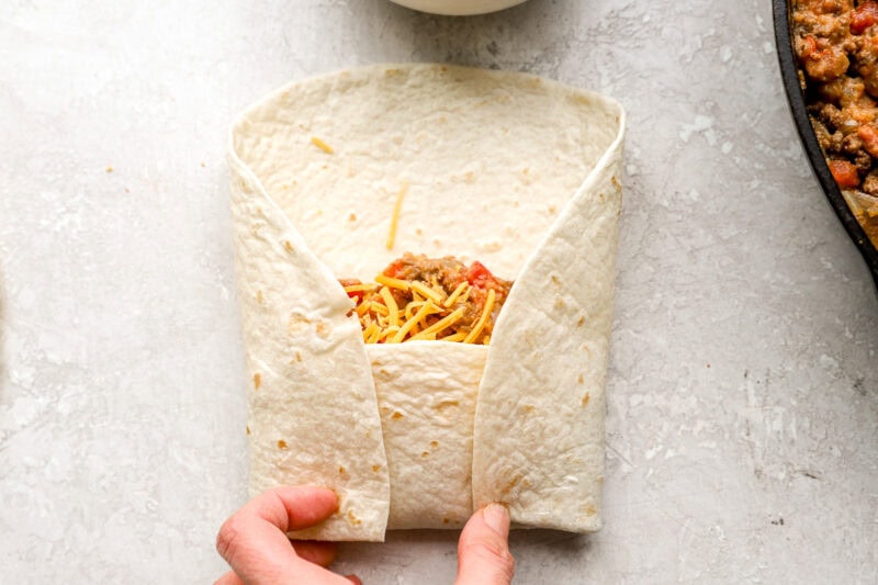 Hands folding a tortilla around the beef and cheese filling, showing the burrito wrapping technique.