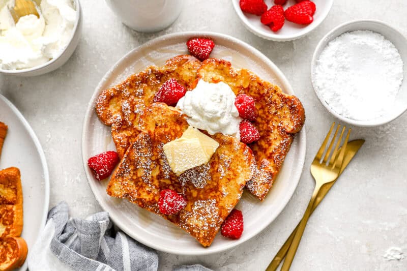 Fully plated French toast with raspberries, whipped cream, butter, and powdered sugar, surrounded by extra toppings and gold utensils.