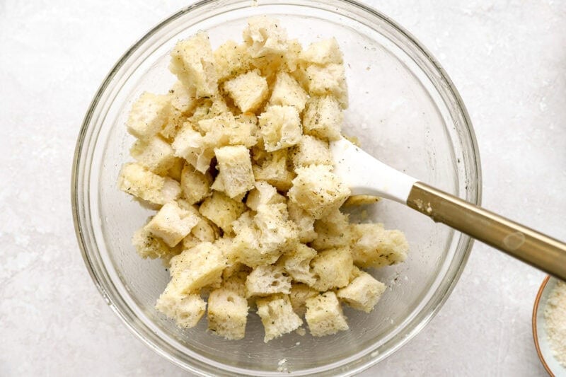 Cubed bread tossed with the seasoned garlic butter mixture in a glass mixing bowl with a white spatula.