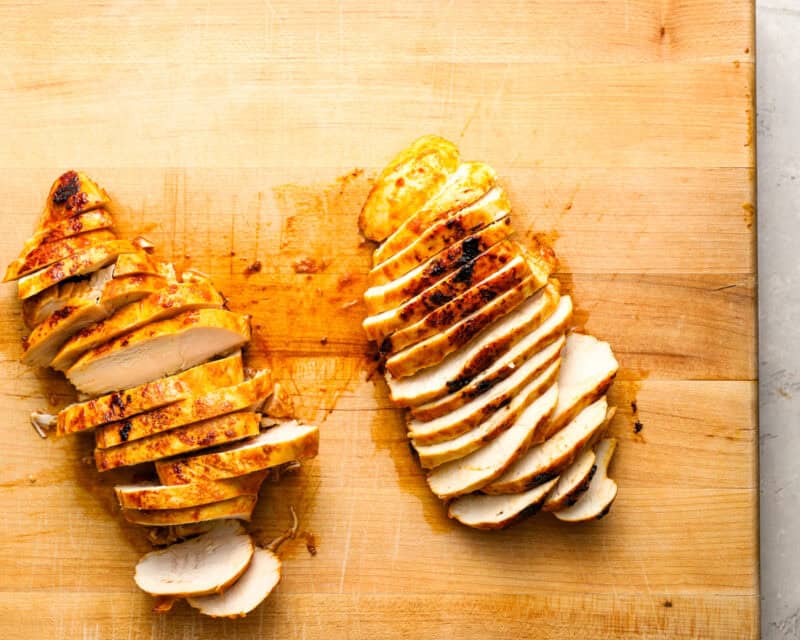 Sliced cooked chicken breasts on a wooden cutting board, showing juicy texture and slightly charred edges.