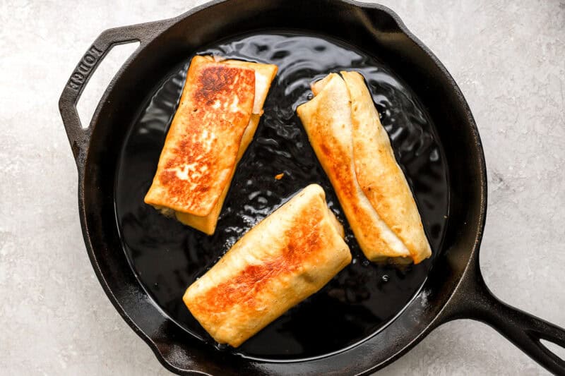 Three chimichangas frying in a cast iron skillet until golden brown and crispy.