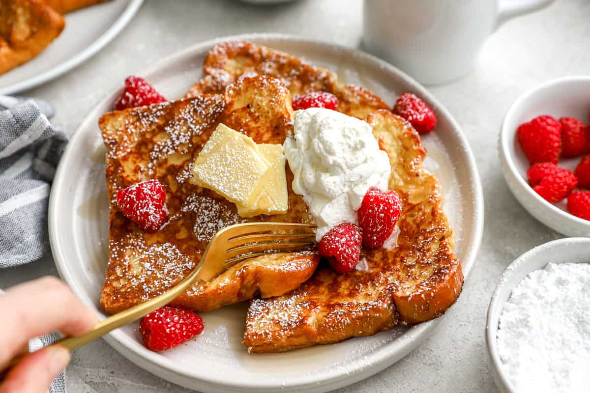 Fork cutting into a slice of brioche French toast topped with whipped cream, butter, powdered sugar, and raspberries.