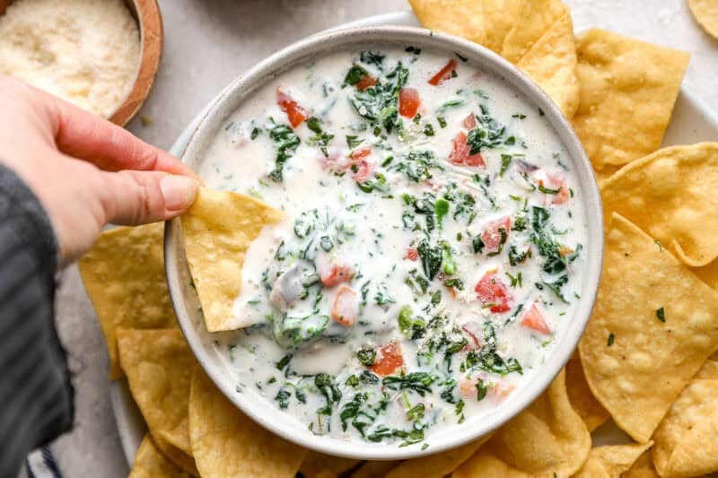 Hand dipping a tortilla chip into a bowl of creamy spinach and pico de gallo cheese dip, surrounded by more chips.