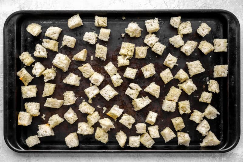 Seasoned bread cubes spread out evenly on a dark baking sheet before baking.